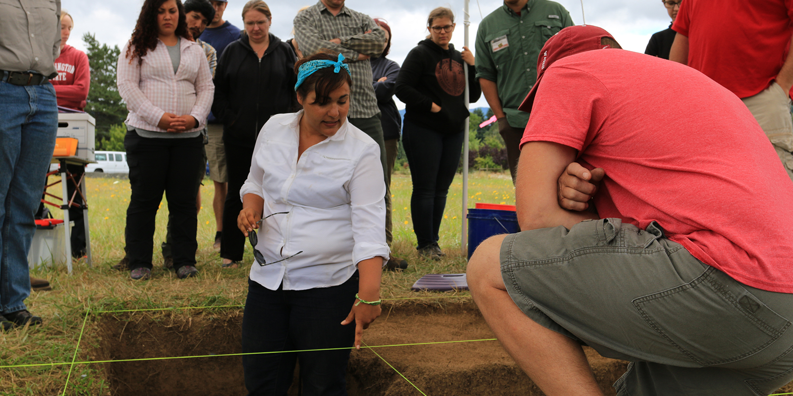 A woman stands in a pit pointing out an archaeology dig to other people